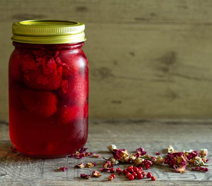 Jar of pickled strawberries next to dried rose petals and pink peppercorns