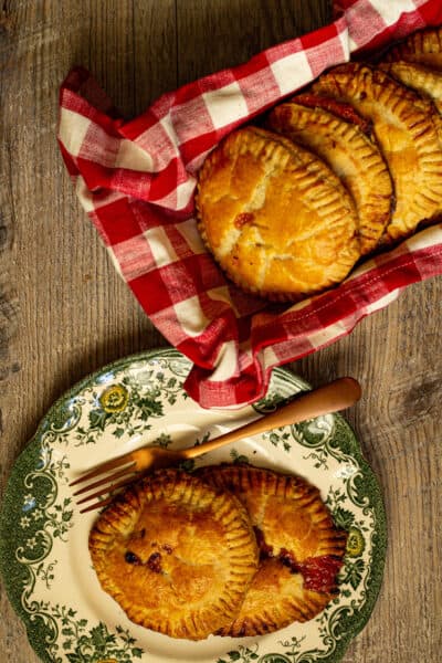 Hand pies on a plate next to hand pies in a dish wrapped in a red and white checked towel