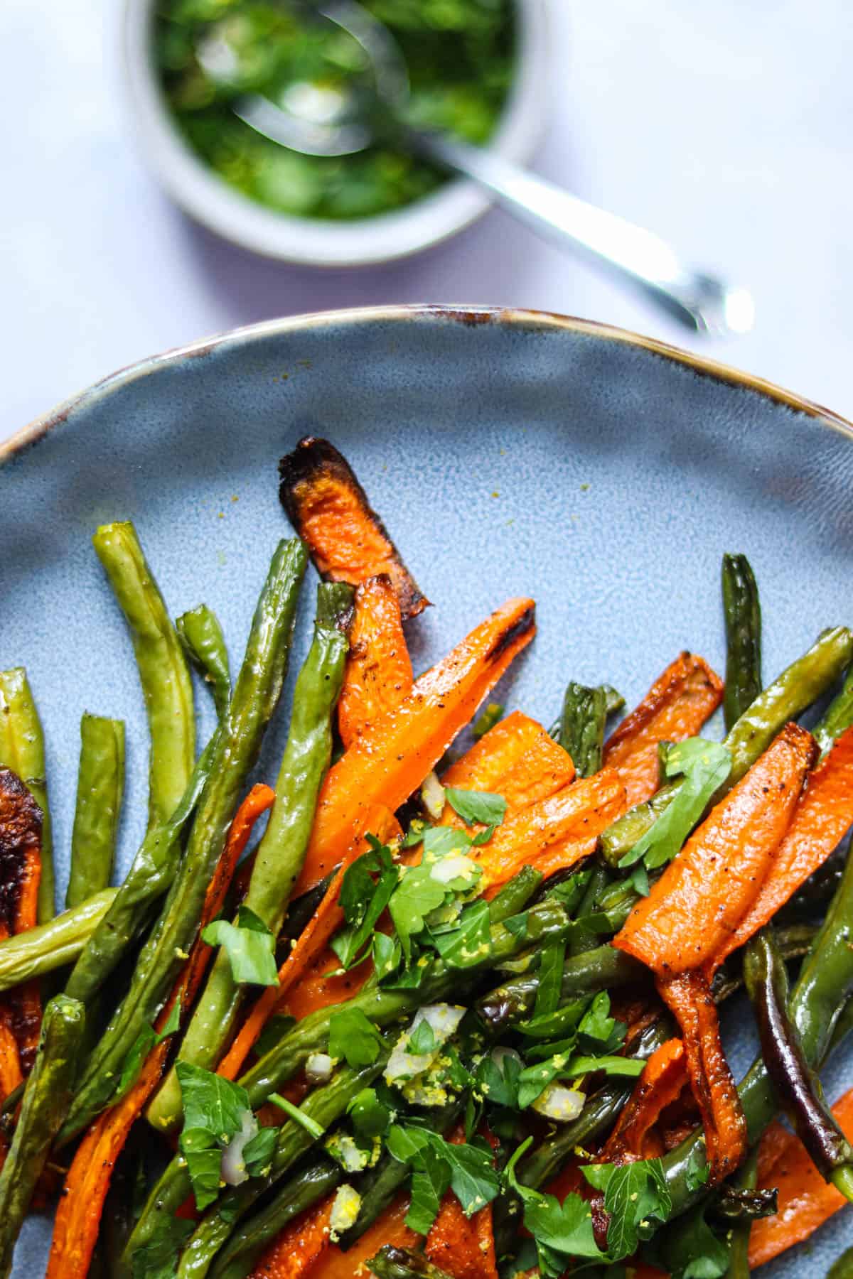 close up photo of roasted green beans and carrots on a blue plate topped with shallot gremolata.