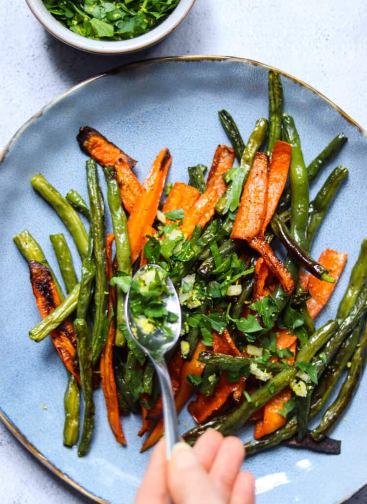 spooning shallot gremolata onto a plate of roasted green beans ad carrots.