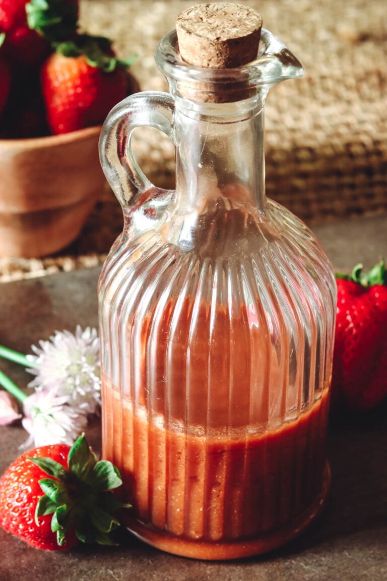 Strawberry balsamic vinaigrette in a glass bottle with a cork next to chive flowers and fresh strawberries.