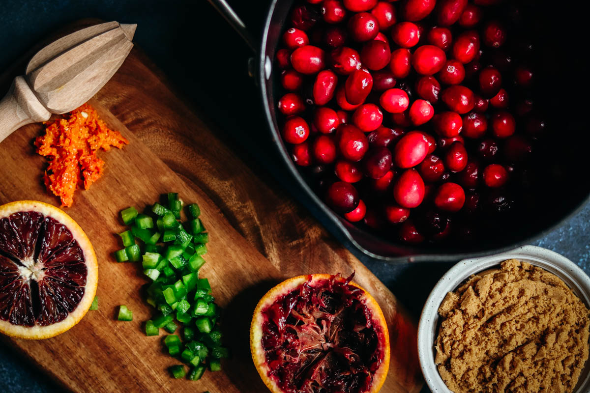 Pot of cranberries and juiced blood orange with chopped jalapeno on a cutting board.