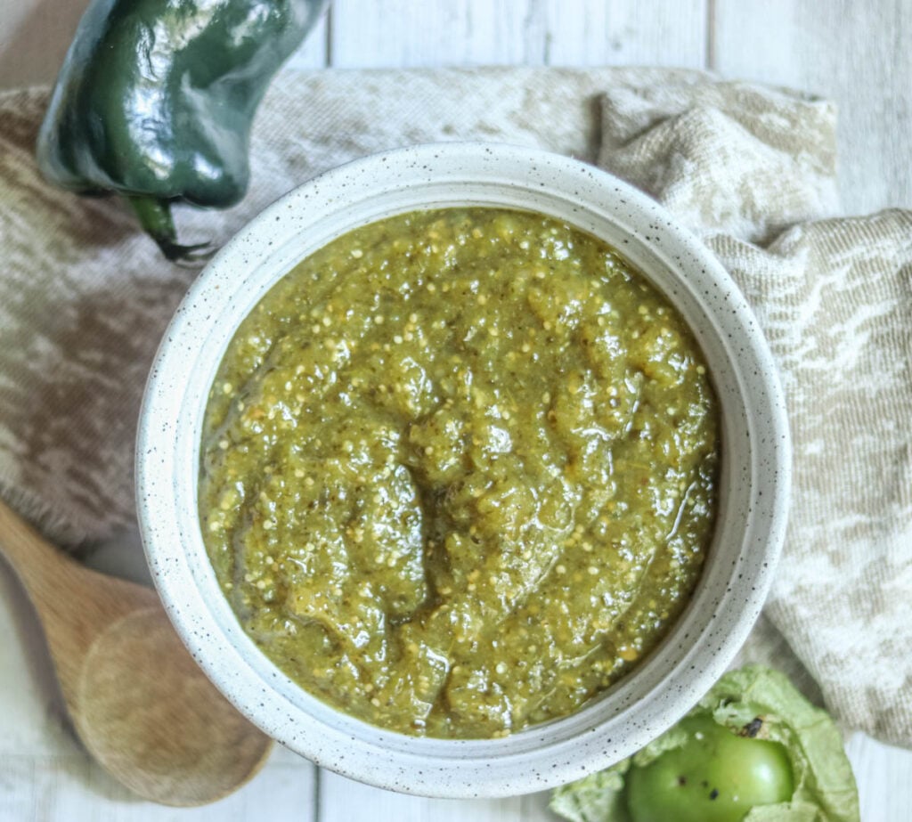 Green Chile salsa in a bowl with a wooden spoon next to it sitting on a napkin.